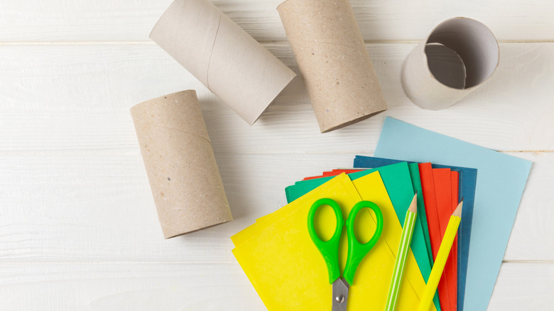 Empty toilet paper rolls, scissors, pencils, and colored paper sheets on a wooden worktable.