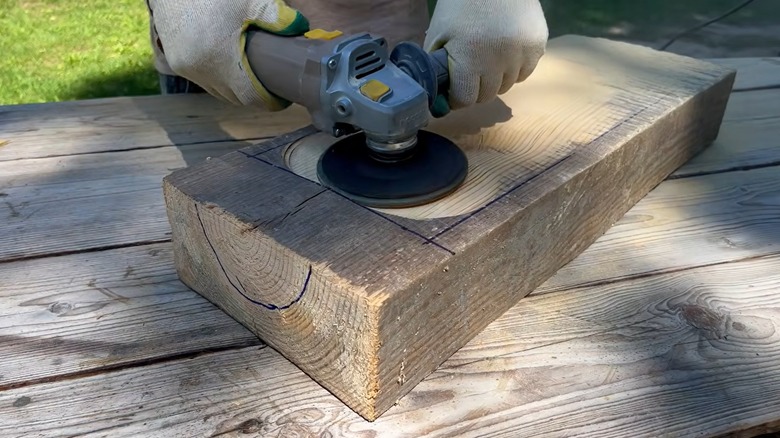 A person sands down a short, thick piece of lumber using an angle grinder.