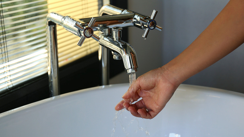 A hand held under a running bathtub faucet in a bathroom.