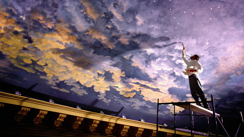 A man stands on scaffolding to paint the sky on a large ceiling.