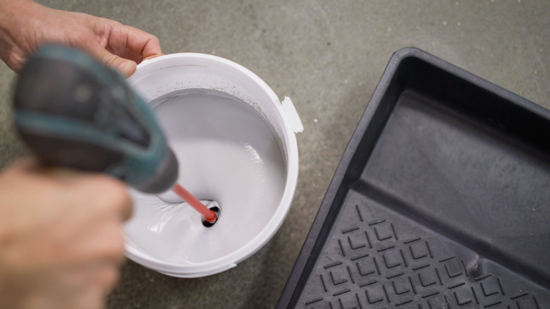 A person mixes paint in a gallon bucket using a drill with a mixer attachment