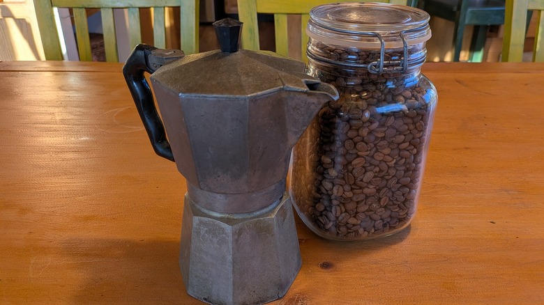 A moka pot and a jar of coffee beans sit on a wooden table.