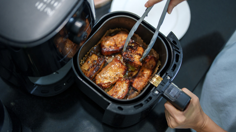 Person using tongs to lift cooked meat from the basket of a black air fryer.