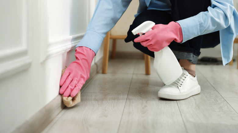 Close-up of a person holding a bottle of cleaning product while wiping a baseboard