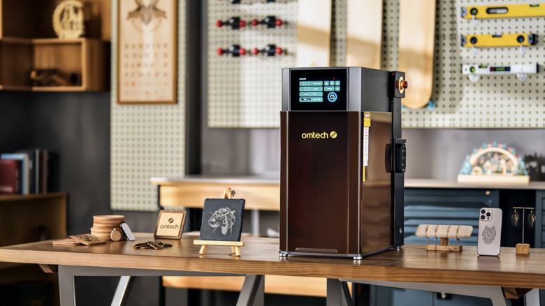 OMTech Solis Duo Dual Laser Engraver on table in workshop, surrounded by finished products like earrings, phone cases, stone display art, and keychains