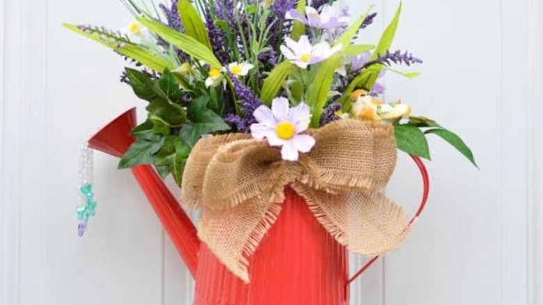 A red watering can is decorated with a burlap bow, artificial green and purple florals, and strands of beads spilling from the spout.