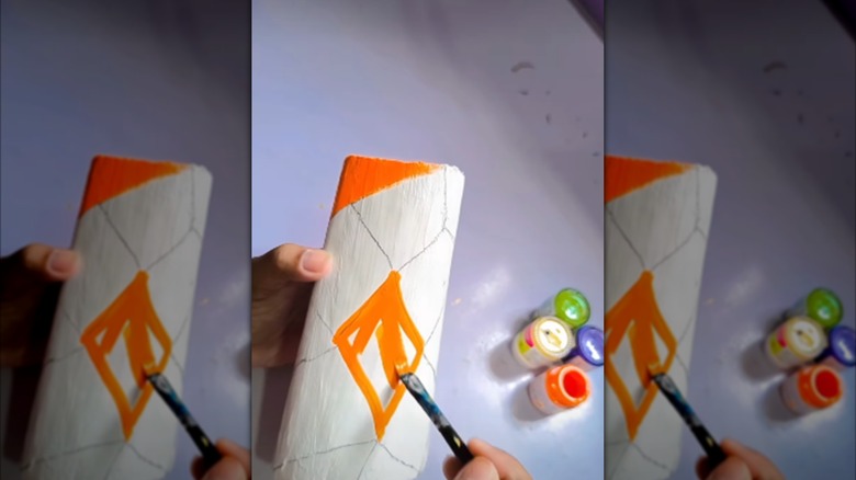 A person paints orange diamond shapes onto a painted empty shampoo bottle with a paintbrush.