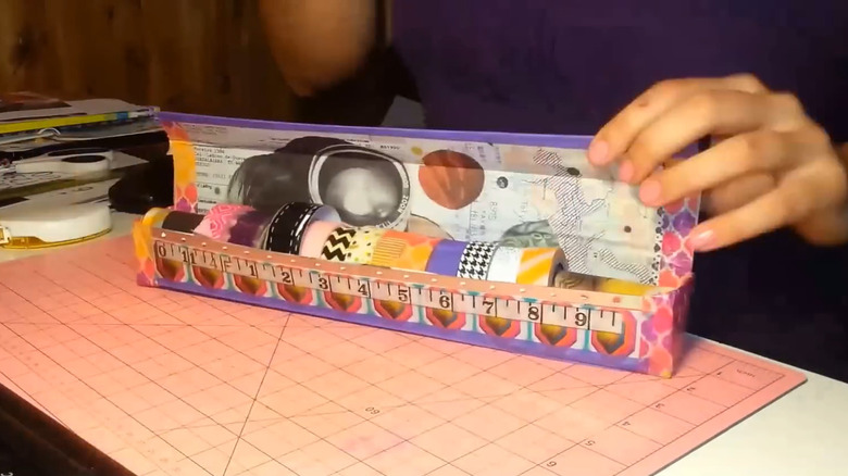 A hand propping a DIY washi tape dispenser box open on a table