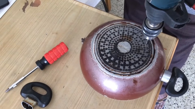 A person drilling holes into an old frying pan on a wooden work bench