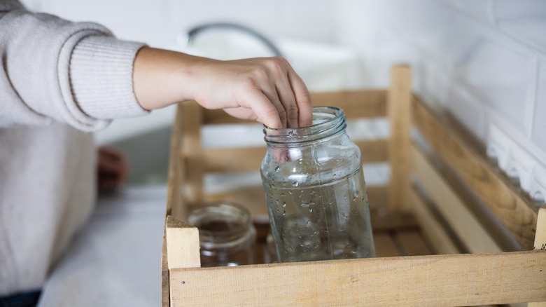 A person puts a recently washed mason jar into a wooden crate on a kitchen countertop.