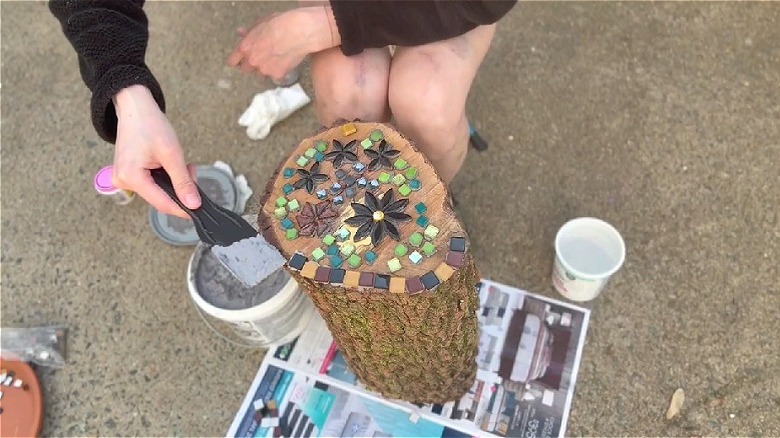 A person using thin set mortar on a spatula to stick mosaic tiles on a cut tree stump.