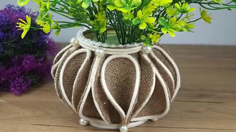 A vase made of a metal can covered in burlap sits on a wooden tabletop nearby some purple flowers and is filled with yellow and green faux flowers.