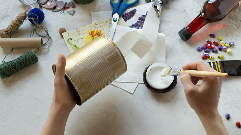 A pair of hands paint glue onto a tin can above a table filled with craft supplies.