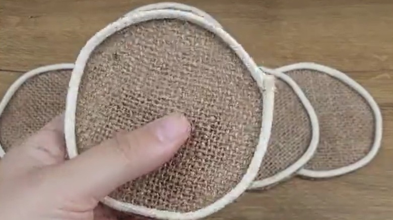 A hand holds circles of burlap edged with white cord above a wooden tabletop.