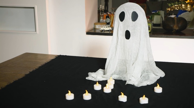 Floating ghosts made out of cheese cloth used as Halloween decor over a wooden table covered with a black towel and fake candles.
