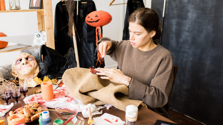 Serious busy young woman in brown sweater sitting at desk full of Halloween stuff and tools and adding colorful autumn leaves while sewing burlap in workshop.
