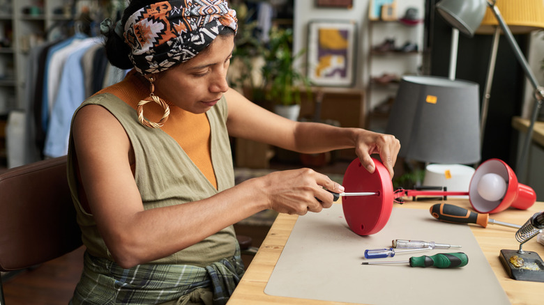 A woman unscrews the bottom off a red desk lamp at her workstation.