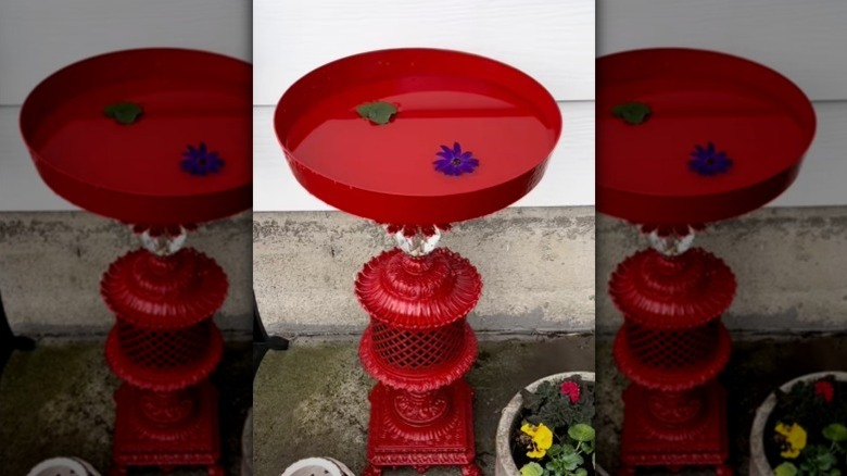 A red-painted bird bath made from an antique baroque lamp base and a large metal dish.