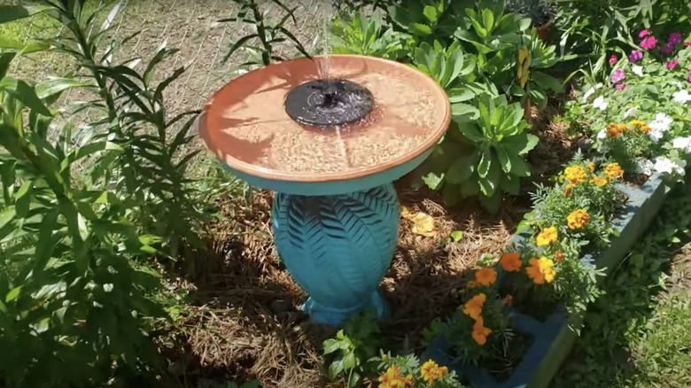 A fountain bird bath made from a ceramic lamp base and a platter stands in a garden bed.