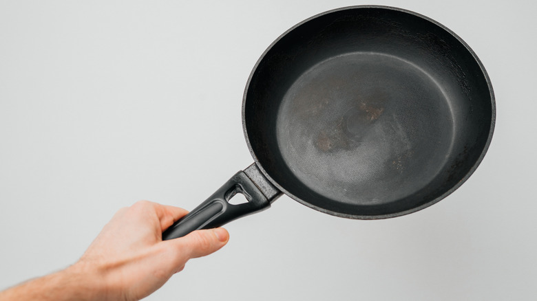 A hand holds an old non-stick frying pan against a white wall.