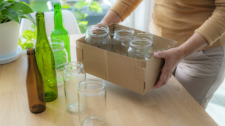 Woman placing cardboard box full of mason jars onto table where other colored glass bottles are placed