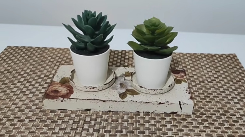 A painted and embellished plant stand made of two jar lids glued onto a wooden block with two mini potted houseplants.