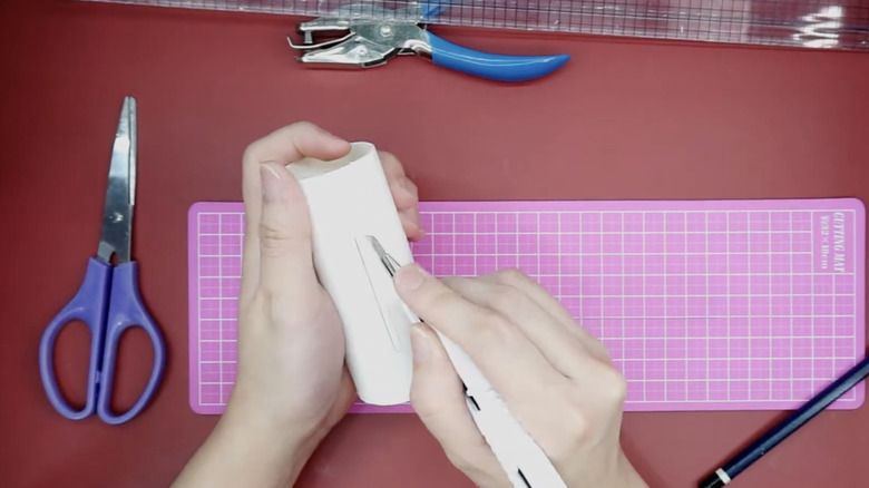 A person uses a scalpel to cut a rectangle out of a toilet paper tube.