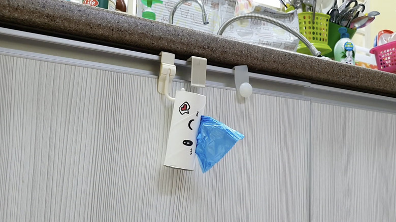 A trash bag dispenser made from a toilet paper tube hangs on a wall with a blue trash bag sticking out of it.