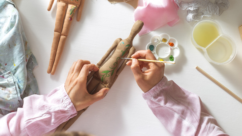 A woman's hands paint a doll on a table covered in craft supplies.