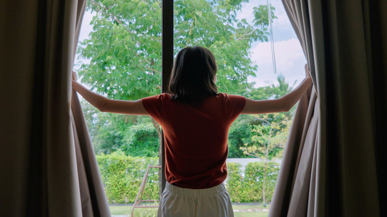 Woman in orange top and white pants holds open heavy drapes and looks out window.