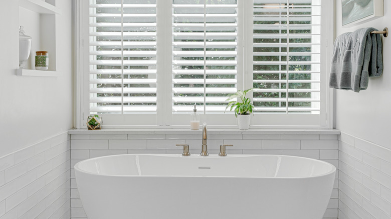 A bathtub under a window with plantation shutters.