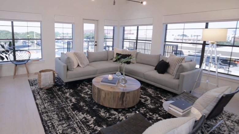 Two walls of windows sit behind an L-shaped gray couch that has a round coffee table in front of it.