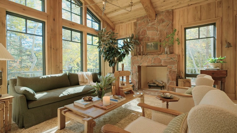 A wood paneled living room with green and white seating areas, wooden furniture, large windows, a stone fireplace, and several plants.