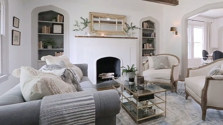 A white and gray living space with recessed bookcases on either side of the fireplace, wooden beams on the ceiling and an arched entryway to the next room.
