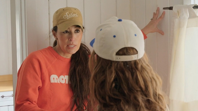 Joanna Gaines wearing an orange sweatshirt and putting hand on wall talking to another woman whose back is to camera