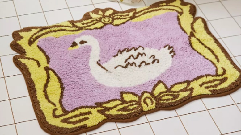 A bathmat decorated with a swan against a pink background and brown and yellow frame detail