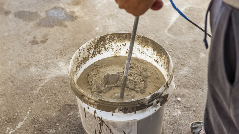 Person mixing concrete powder with water in a 5 galon bucket ona. paved surface