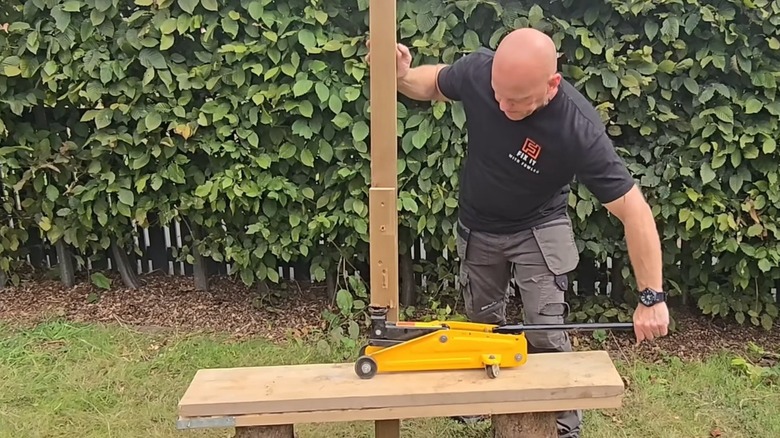 A man uses a car jack to pop a fence post out of the ground.