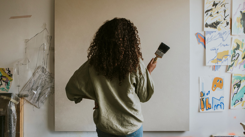 A back view of a woman with long dark hair and a paintbrush inspecting a blank canvas