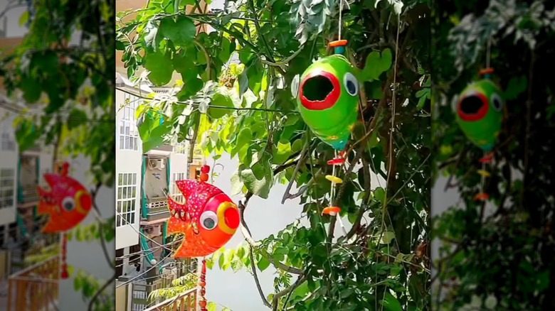 Painted red, orange, and green fish made from plastic bottles are hung with string on tree branches.