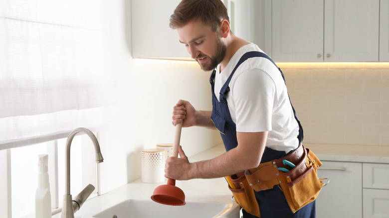 A man holding a plunger tries to unclog sink in a clean, modern kitchen.