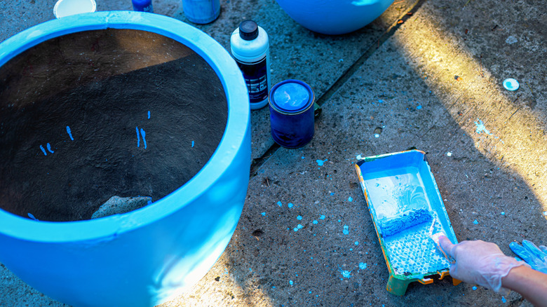 A person working outdoors paints a flower pot blue with a paint roller.