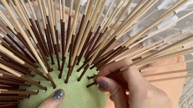 Adding brown skewers to a green styrofoam ball.