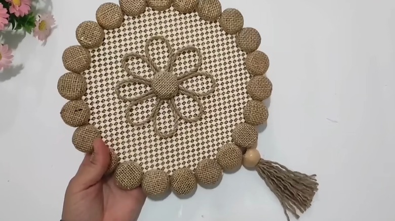 A hand holds some DIY hanging wall art made of hessian, anti-slip mat, twine, a tassel, and a wooden bead.