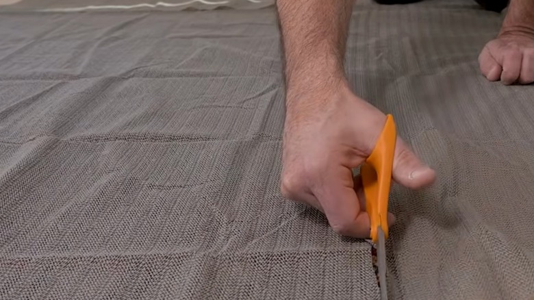 A man cuts through a large piece of anti-slip netting using a pair of orange-handled scissors.
