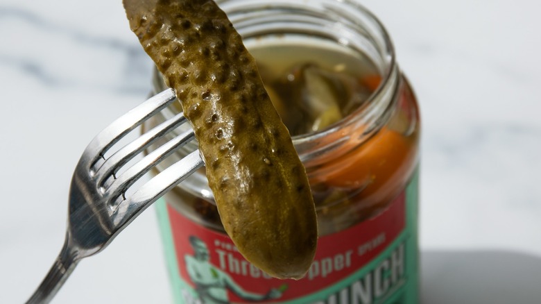 A silver fork holding a pickle in front of an open pickle jar on a white background