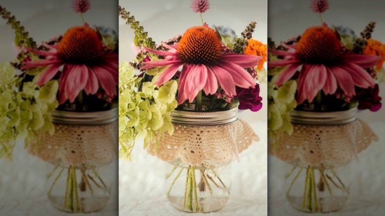 An old glass jar and doily are used to keep a pink coneflower and other flowers in place.
