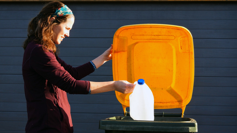 A woman pauses before throwing an empty plastic milk jug into a large outdoor trash can.