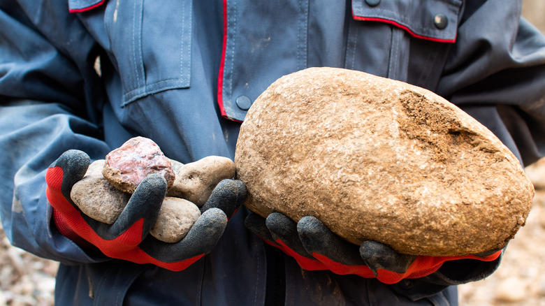 A person wearing gloves holds a large river stone in one hand and a pile of small river stones in the other hand.