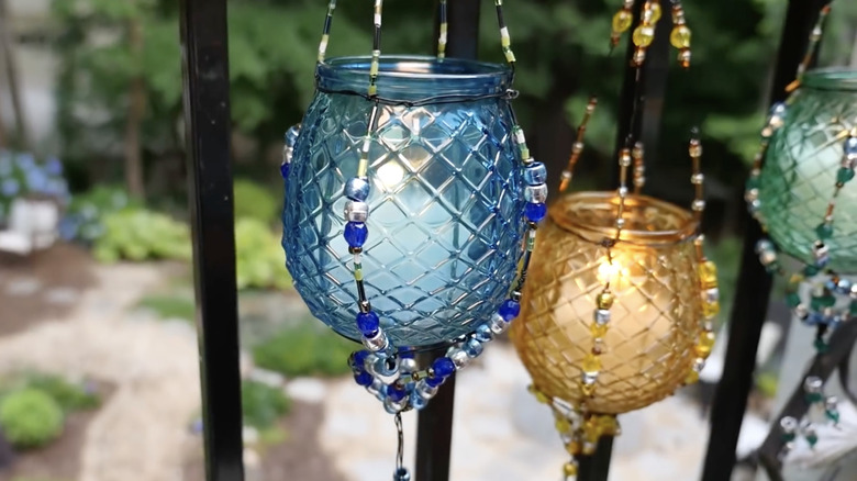 Colorful glass jar lanterns in beaded wire cradles hang from the railings of a balcony with a garden in the background,